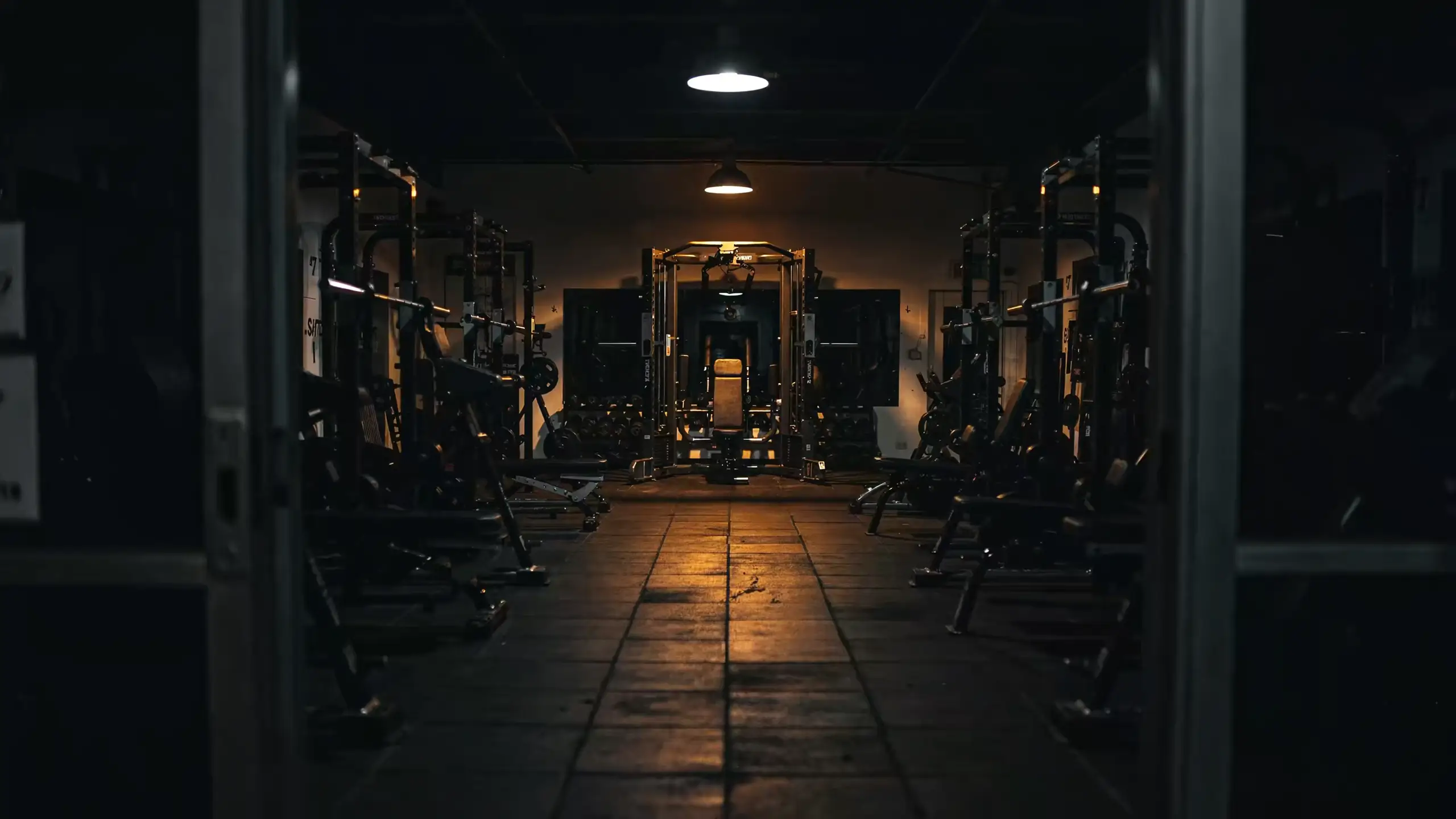 Walking into a dark gym at night, down an aisle of equipment toward a single lit rack at the far end.